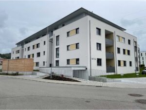 Modernes weißes Mehrfamilienhaus der FÖFA, Hausnummer 14/16, mit Balkonen und Holzverkleidung am Eingangsbereich