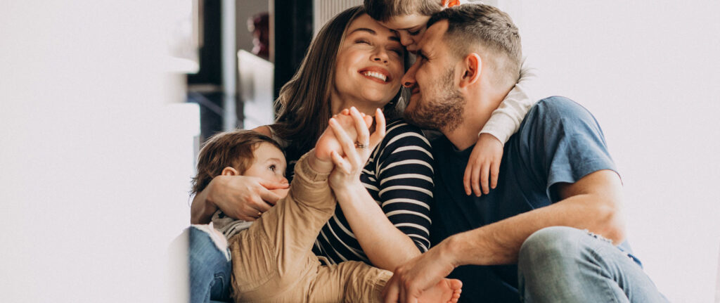 Glückliche Familie mit Kind umarmt sich liebevoll im Wohnzimmer – Hero-Bild der FÖFA Startseite